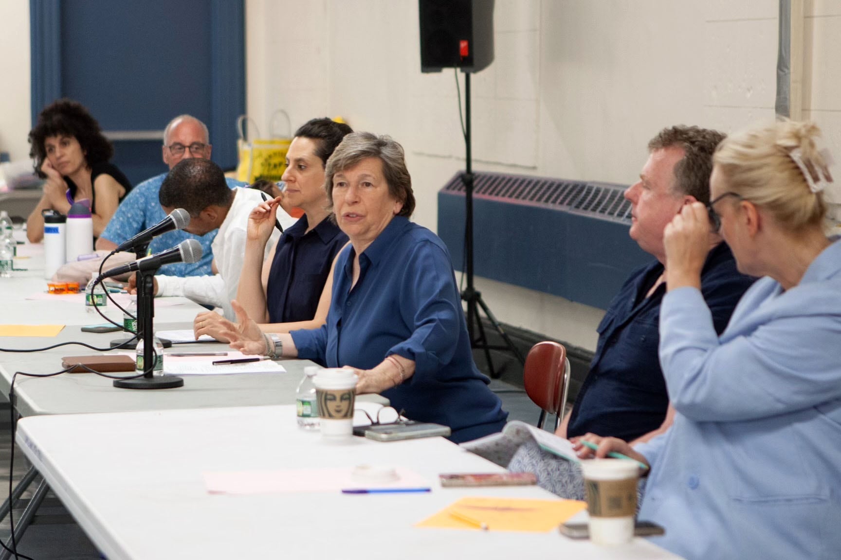A line of adults sit at a white table with a speaker and white wall in the background.