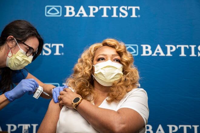 A woman in a mask gets the COVID-19 vaccine in her arm in front of a back drop with the Baptist hospital logo