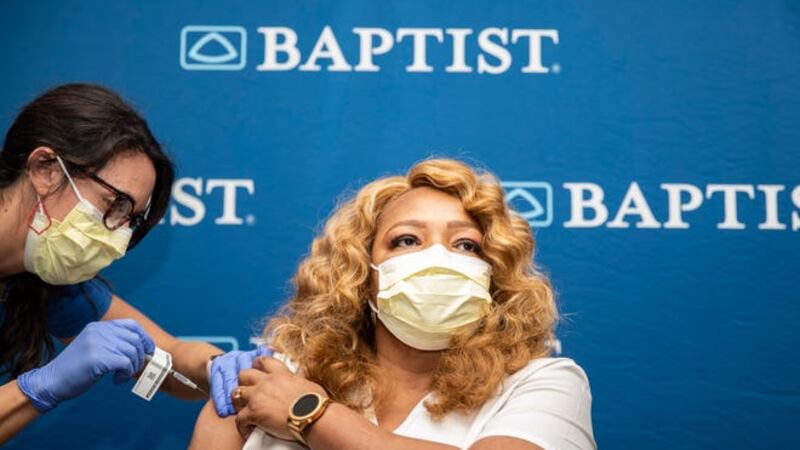 A woman in a mask gets the COVID-19 vaccine in her arm in front of a back drop with the Baptist hospital logo