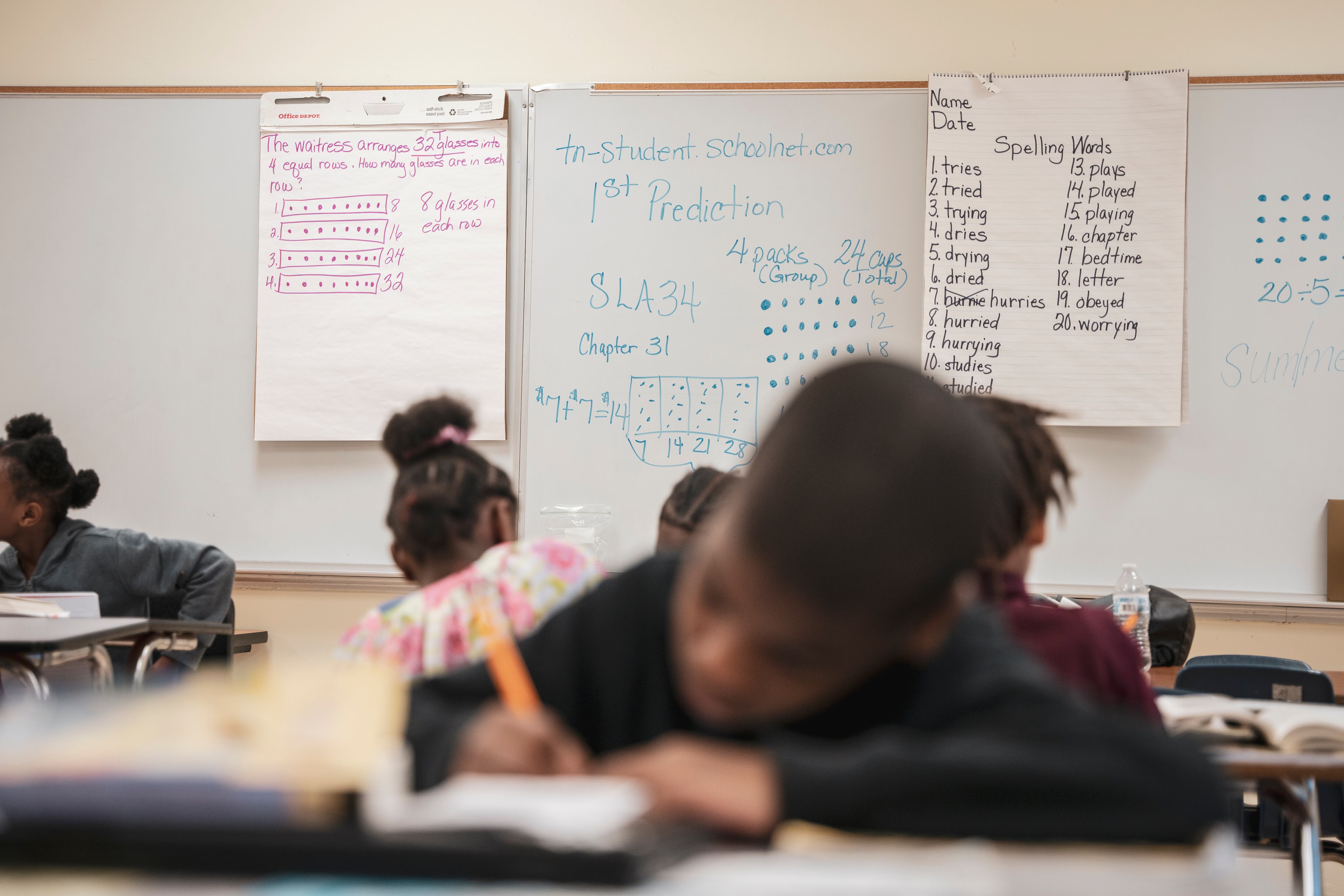A whiteboard with large posters with spelling words and other school work in the background with several children working at desks in the foreground.