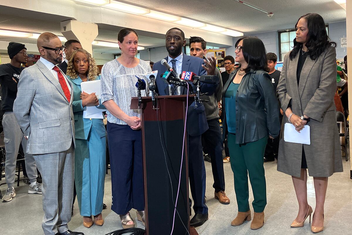 A photograph of a Black man in a suit standing behind a podium speaking into microphones with a large group of people standing around him.