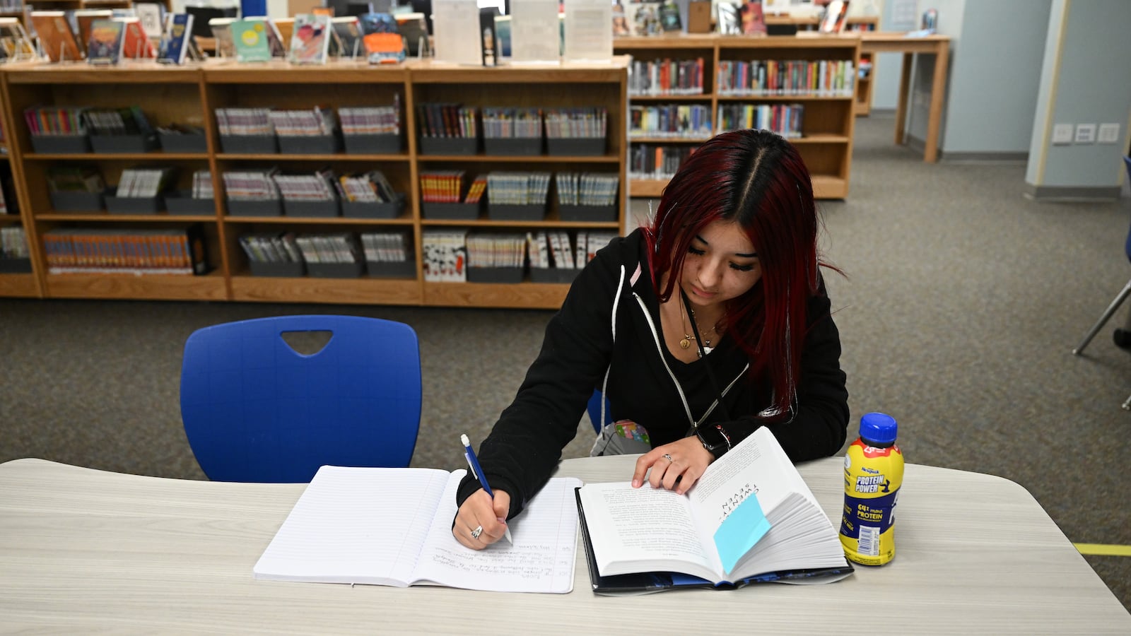 A high school student works on school work with bookshelves in the background.