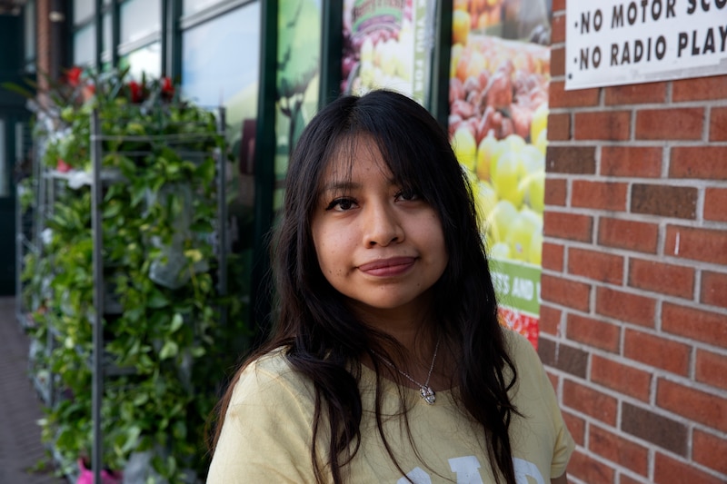 A young person wearing a tan shirt stands in front of a red brick building.