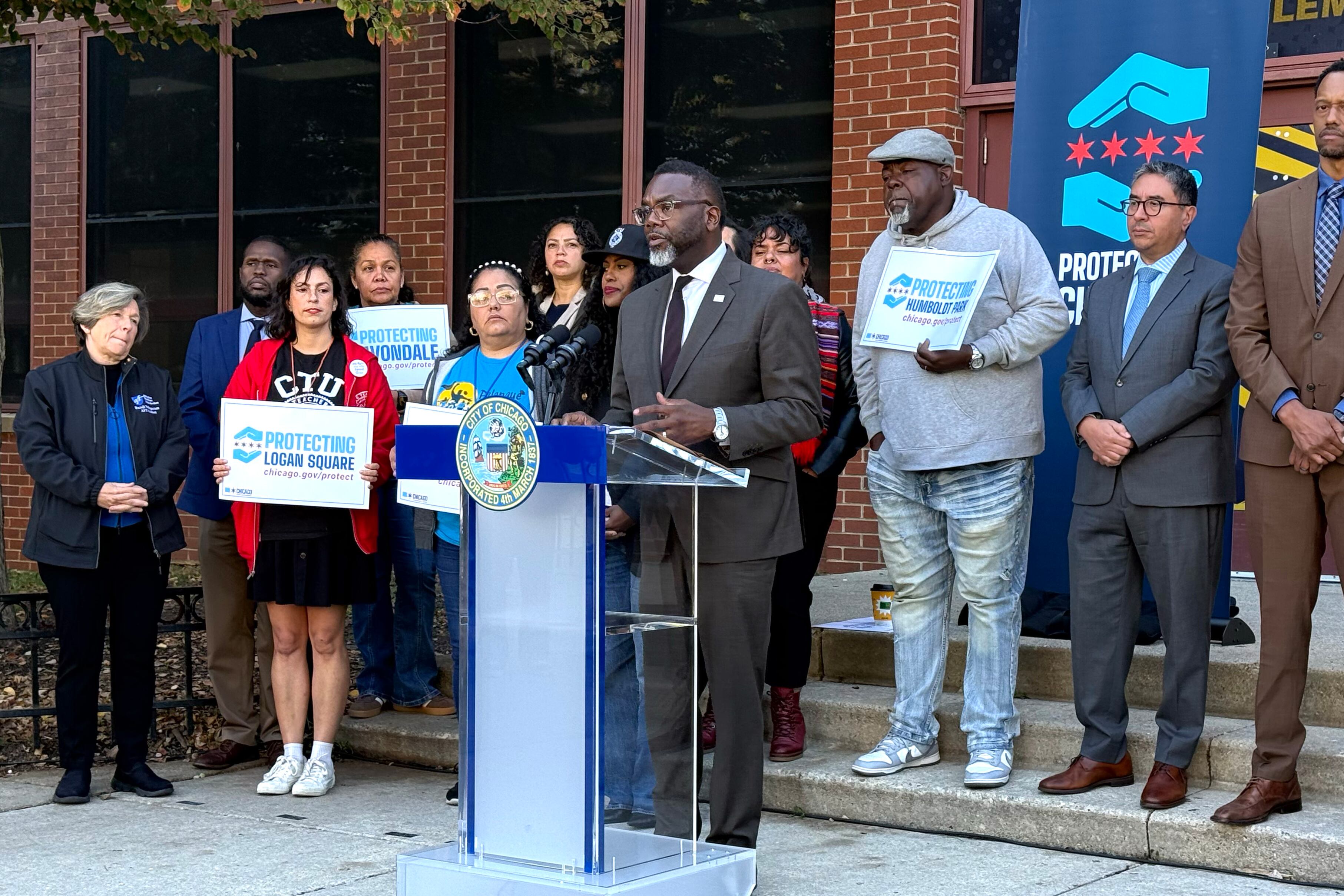 A man in a gray suit speaks while others, some holding signs, stand behind him.