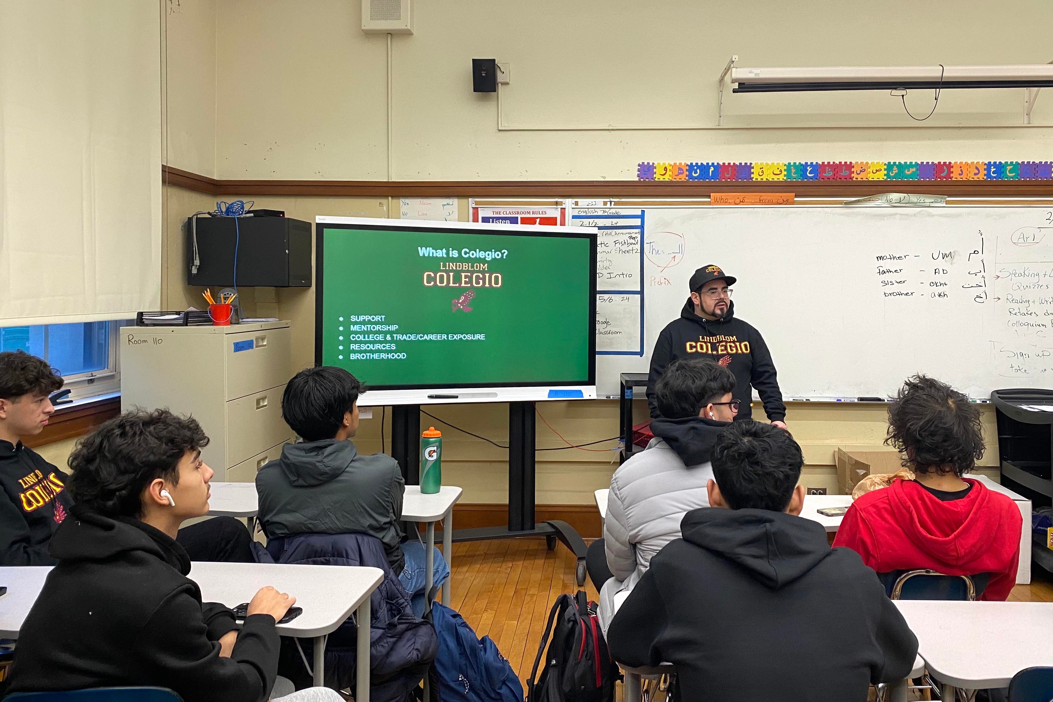 An adult wearing a black hat and sweater stands in front of a class of students at their desks with a white board and a tv screen in the background.