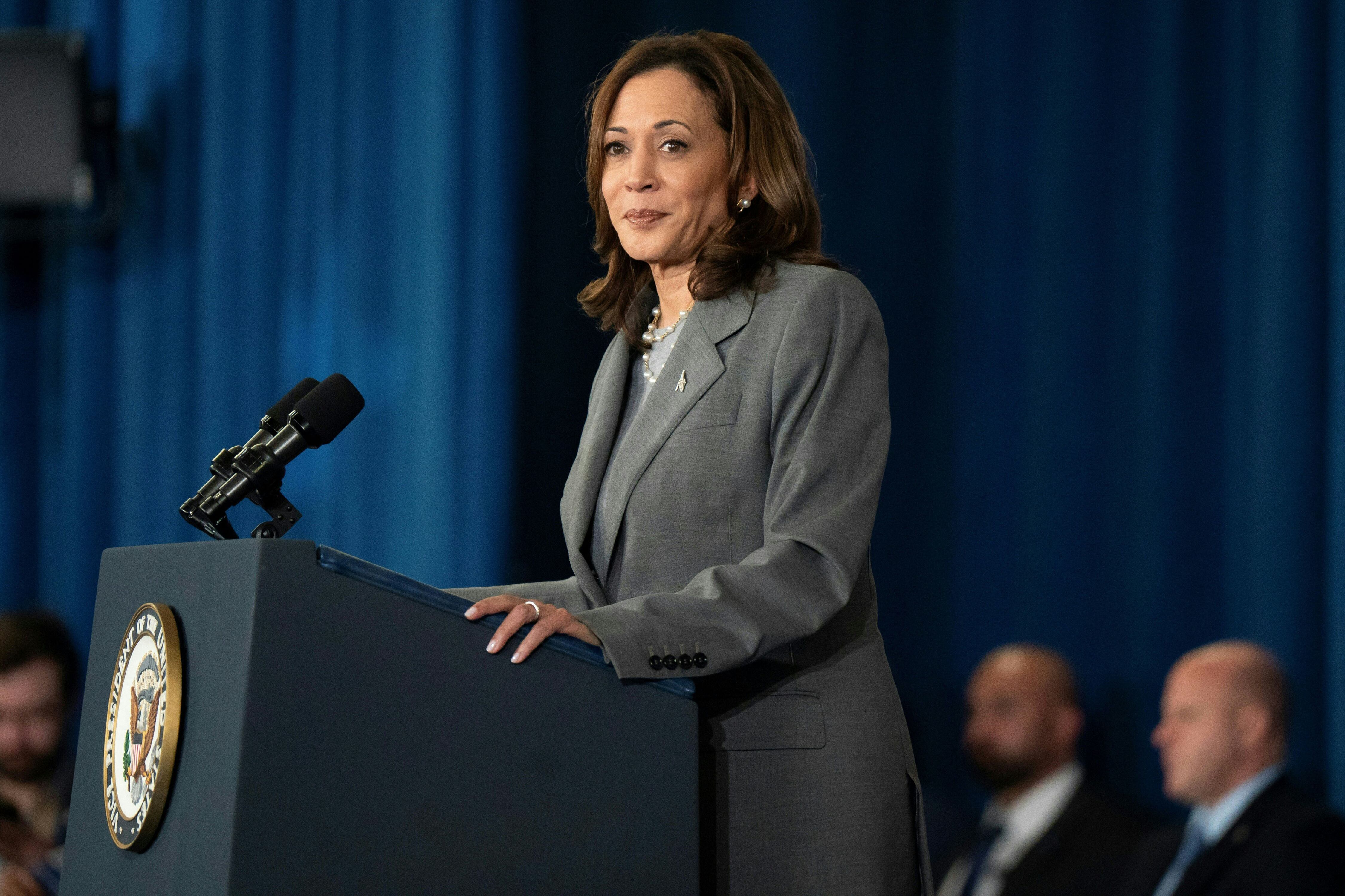 A woman stands at a podium with three men sitting in the background.
