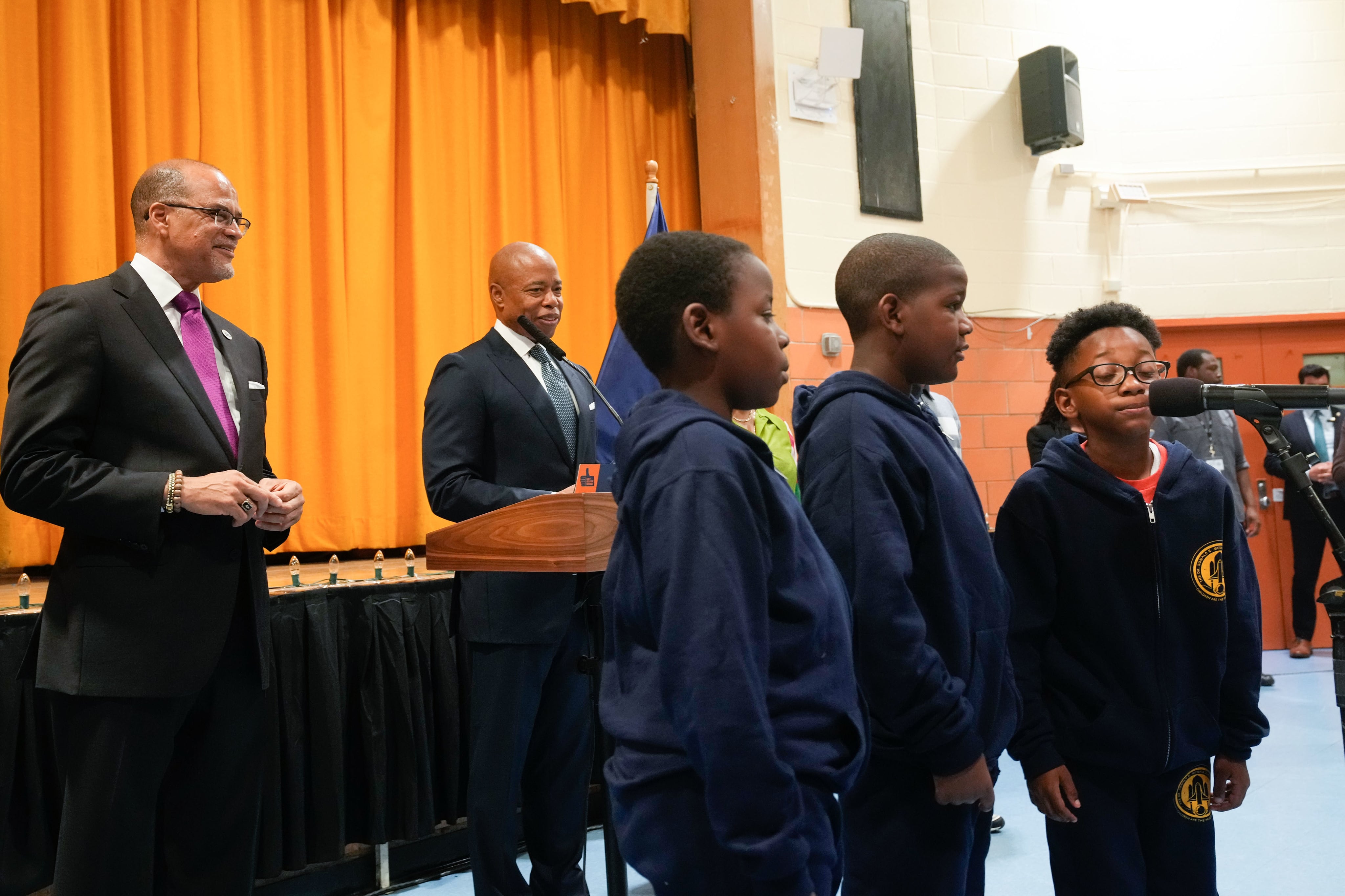 New York schools Chancellor David Banks and New York Mayor Eric Adams stand behind three students who are practicing deep breathing in a room.
