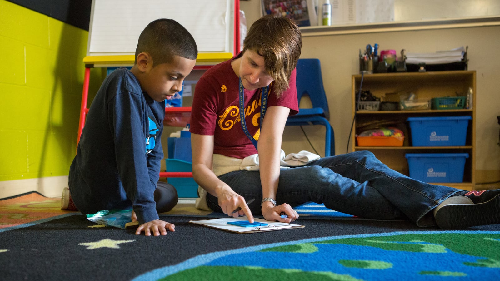 A teacher and a student sit on a rug shaped like the Earth. The teacher is pointing to a piece of paper and the student is looking at it.