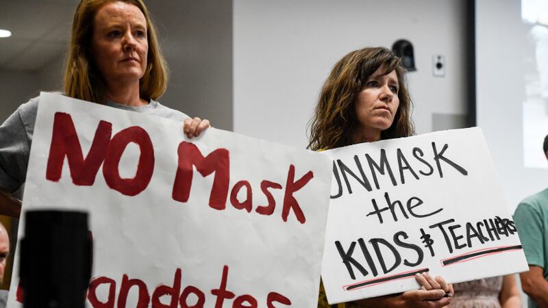 Two women hold signs that say “No mask mandates” and “Unmask the kids and teachers” at a meeting. They have unhappy looks on their faces.