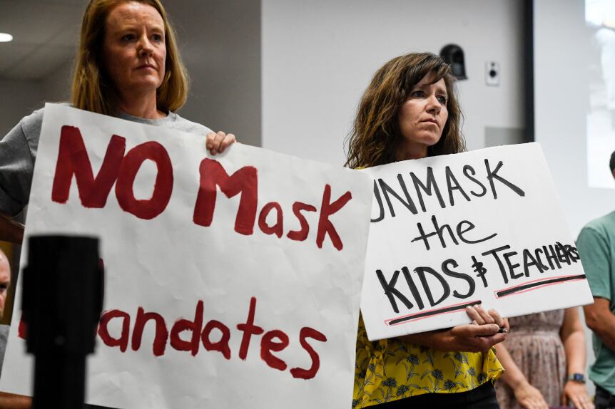 Two women hold signs that say “No mask mandates” and “Unmask the kids and teachers” at a meeting. They have unhappy looks on their faces.