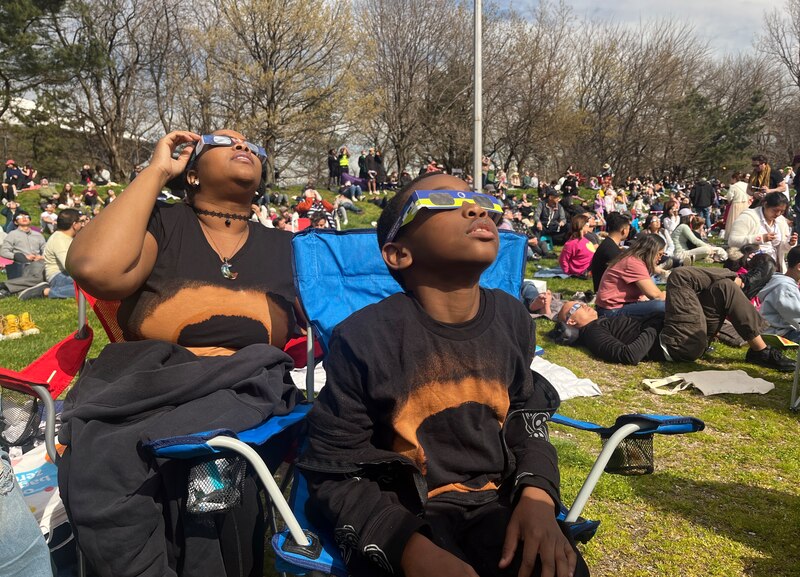 A mom and a 9-yr-old son wear solar eclipse viewing glasses while sitting in lawn chairs on a grassy area with a large group of people sitting behind them.