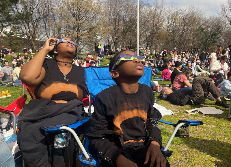A mom and a 9-yr-old son wear solar eclipse viewing glasses while sitting in lawn chairs on a grassy area with a large group of people sitting behind them.