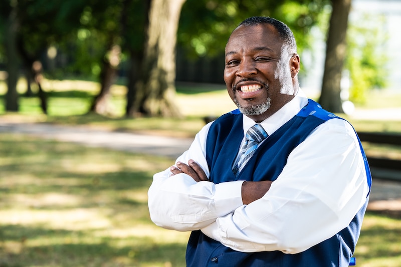 A portrait of a man in a blue vest, white shirt and tie with his arms crossed.