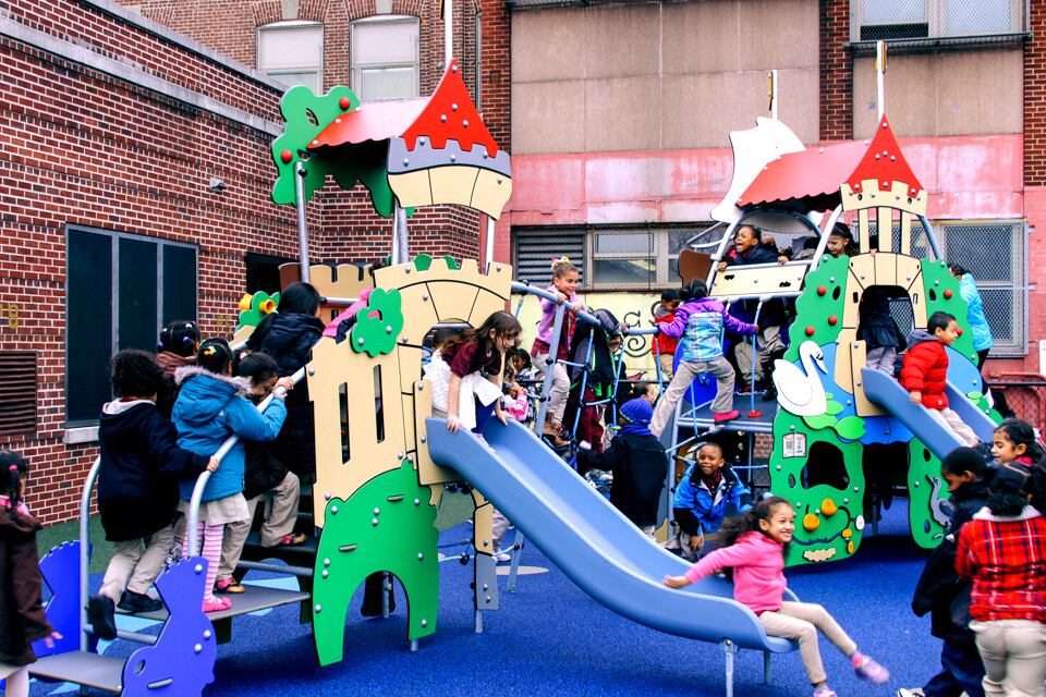 Dozens of children walk up the steps of a yellow, red, and green school playground and slide down blue slides.