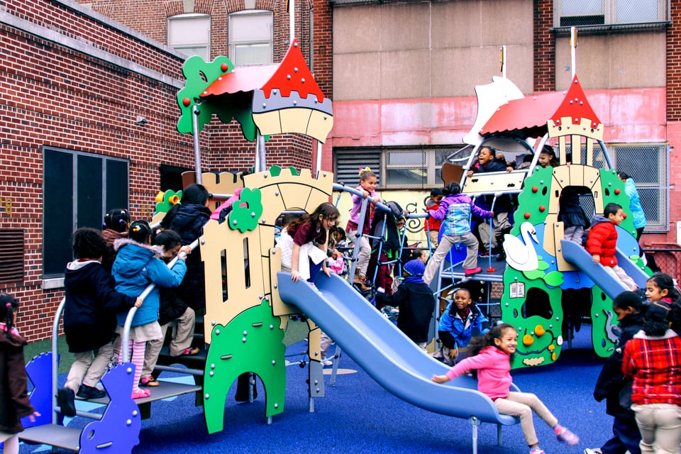 Dozens of children walk up the steps of a yellow, red, and green school playground and slide down blue slides.