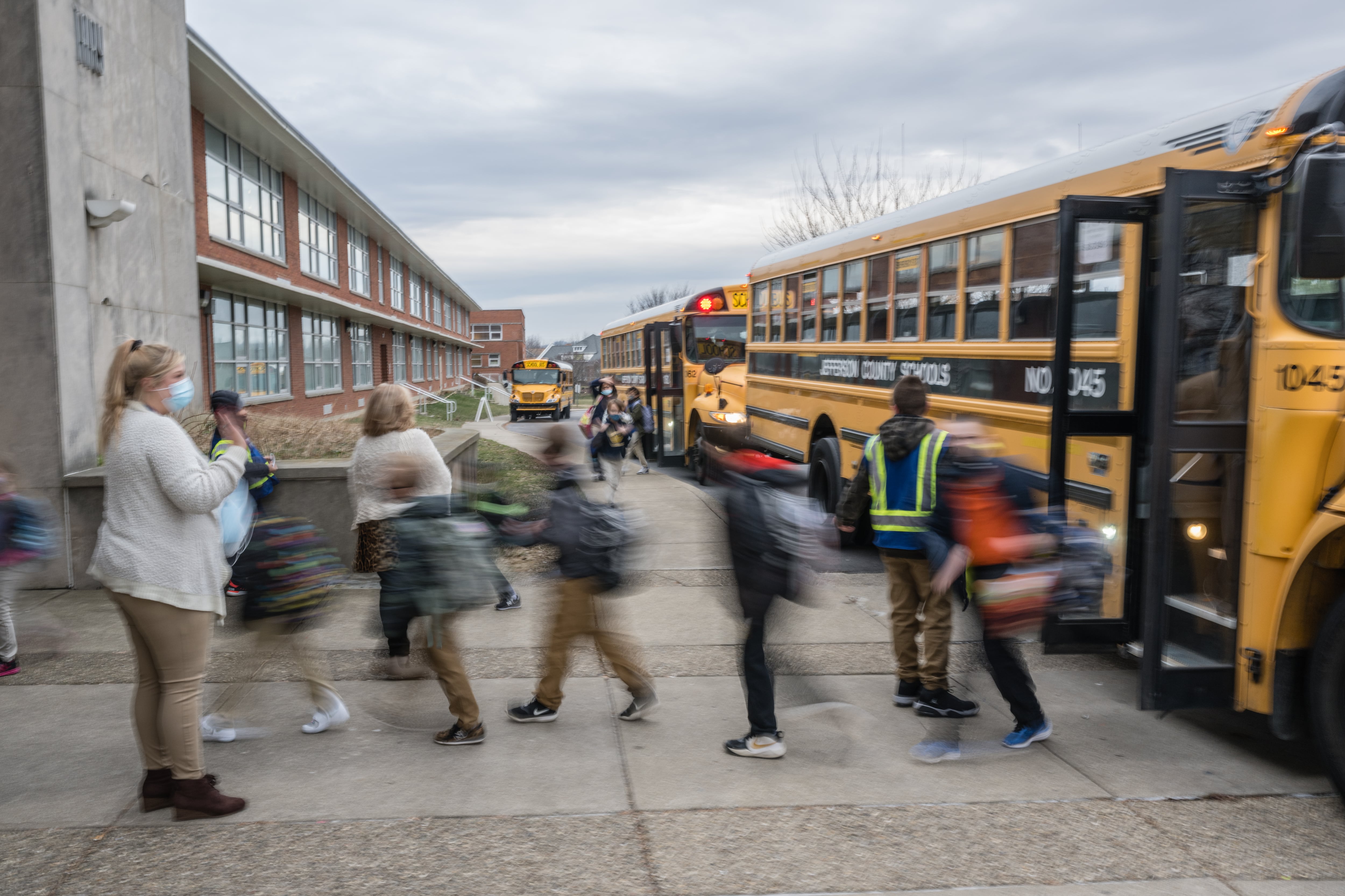 A line of students runs off a school bus and enters a school while a woman in a white sweater supervises.