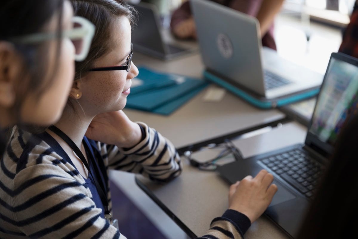 A student works on a laptop with an adult peaking over their shoulder. Other laptops in the background.