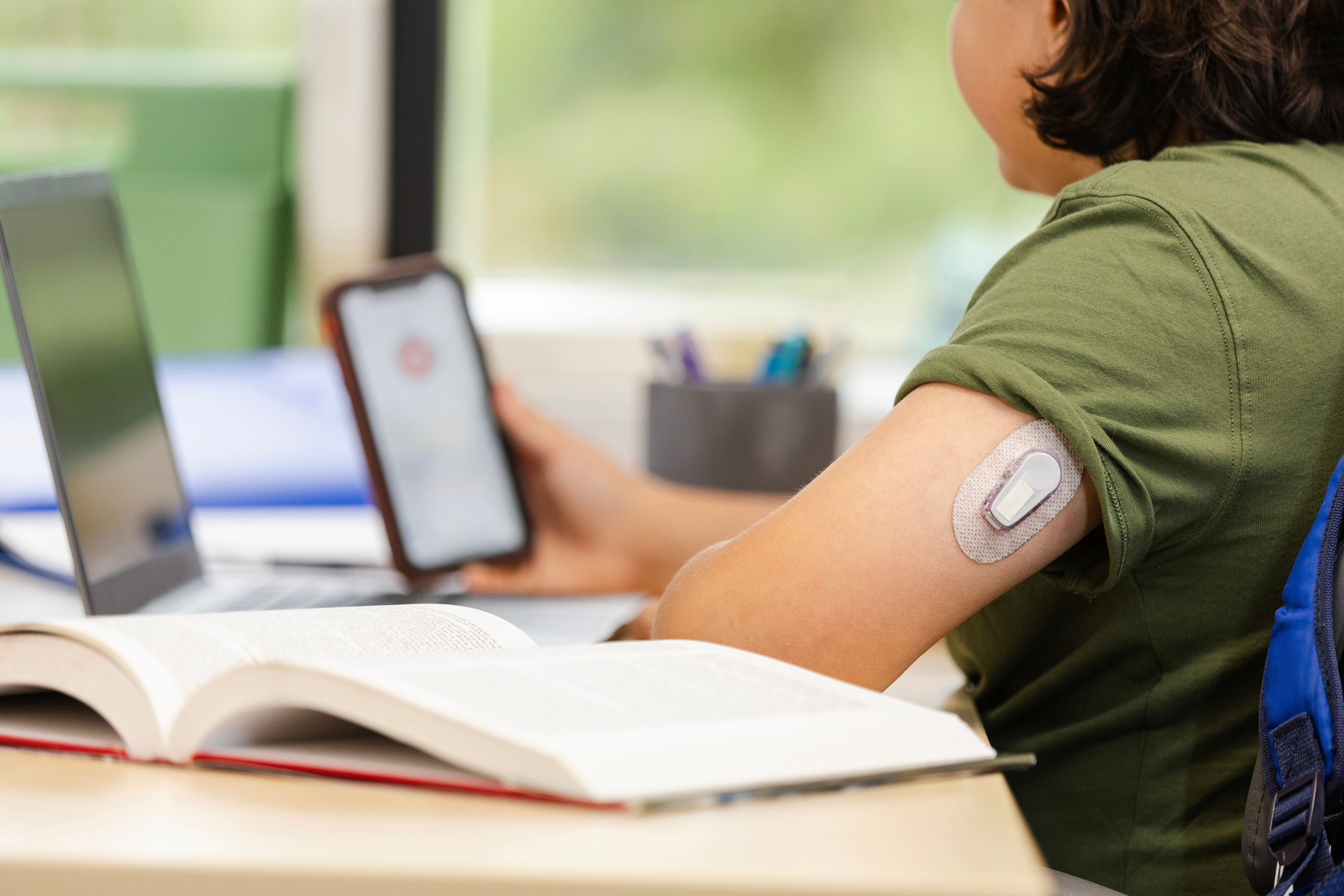 A young student checks their phone at a desk with a text book and laptop while they have a glucose monitor on their arm.