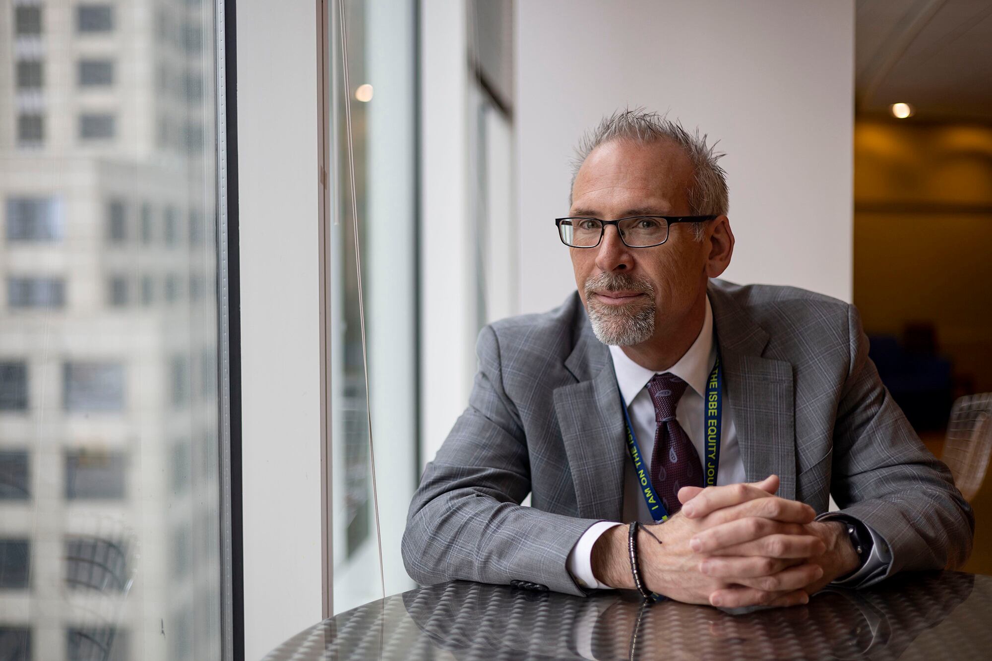 A man in a suit poses for a portrait while sitting at a wooden table near a window.