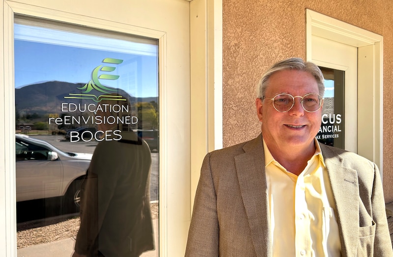A man in a tan jacket and light-colored shirt stands outside a door