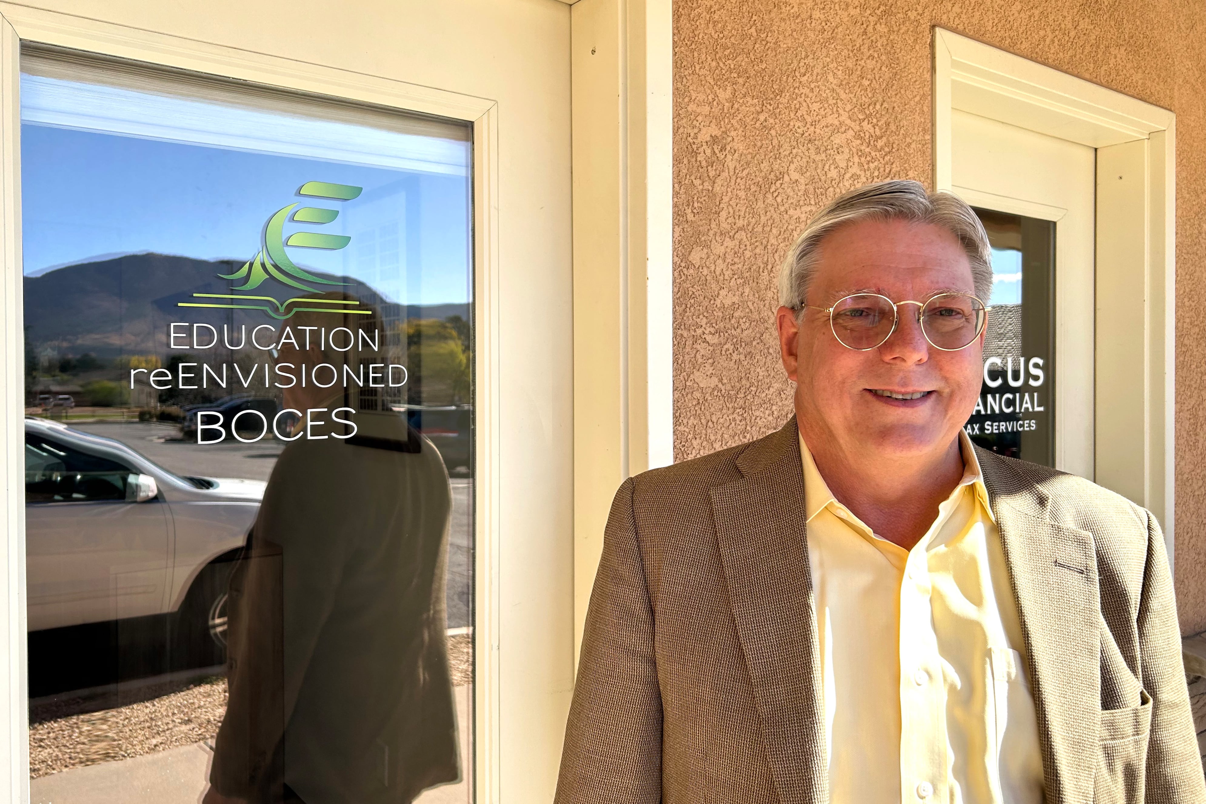 A man in a tan jacket and light-colored shirt stands outside a door