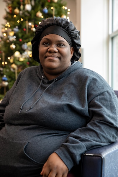 A woman wearing a black hair bonnet and a dark blue sweater, looks at the camera and smiles while a Christmas tree is in the background.