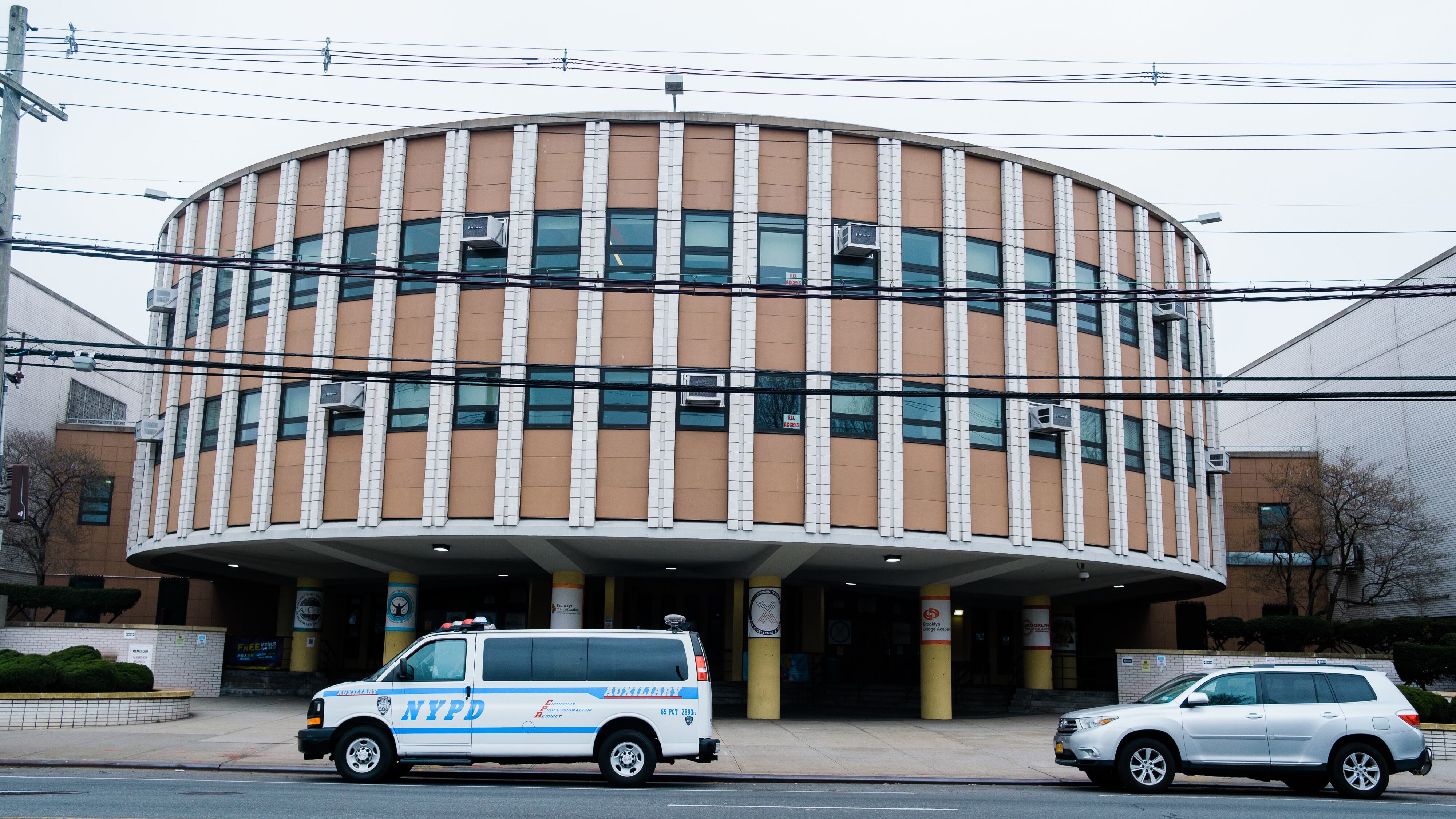 The curved facade of Brooklyn High School for Excellence and Equity in Canarsie. There is an NYPD van parked outside, as well as a standard silver SUV.