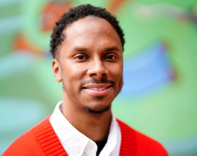A man with short dark hair and wearing a red sweater and white shirt poses for a photo in front of a green background.