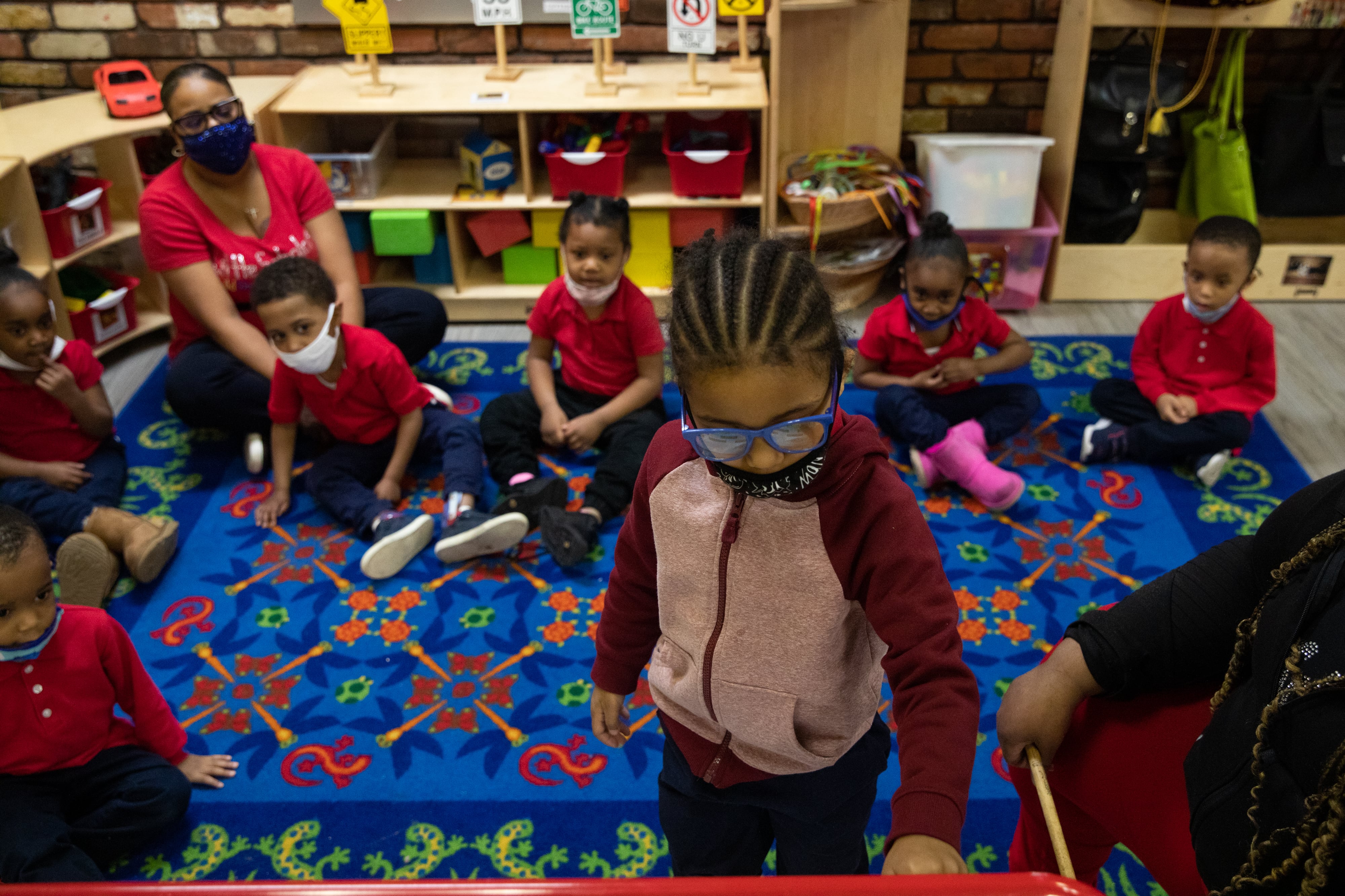 A group of children and a teacher wearing red shirts and dark pants sit on a patterned blue rug wearing masks and watching as a student with cornrows and glasses works on an activity at a whiteboard.