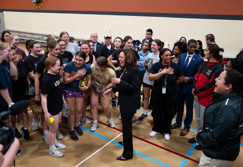 An adult woman stands in front of a crowd of teenage girls in a school gym,.