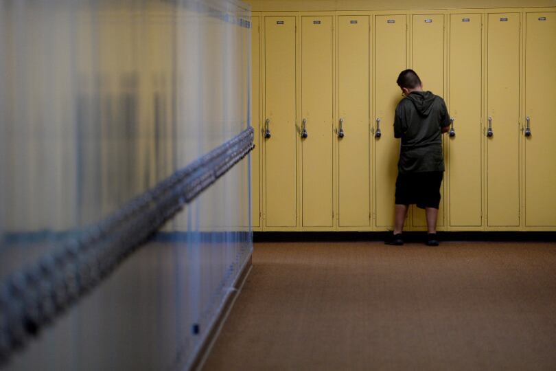 A wide hallway ends in a bank of yellow lockers. A boy who looks about 11 or 12, with short hair and a black T-shirt and shorts, is seen from behind. He’s opening one of the lockers. He’s alone.
