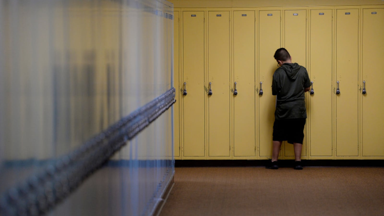 A wide hallway ends in a bank of yellow lockers. A boy who looks about 11 or 12, with short hair and a black T-shirt and shorts, is seen from behind. He’s opening one of the lockers. He’s alone.