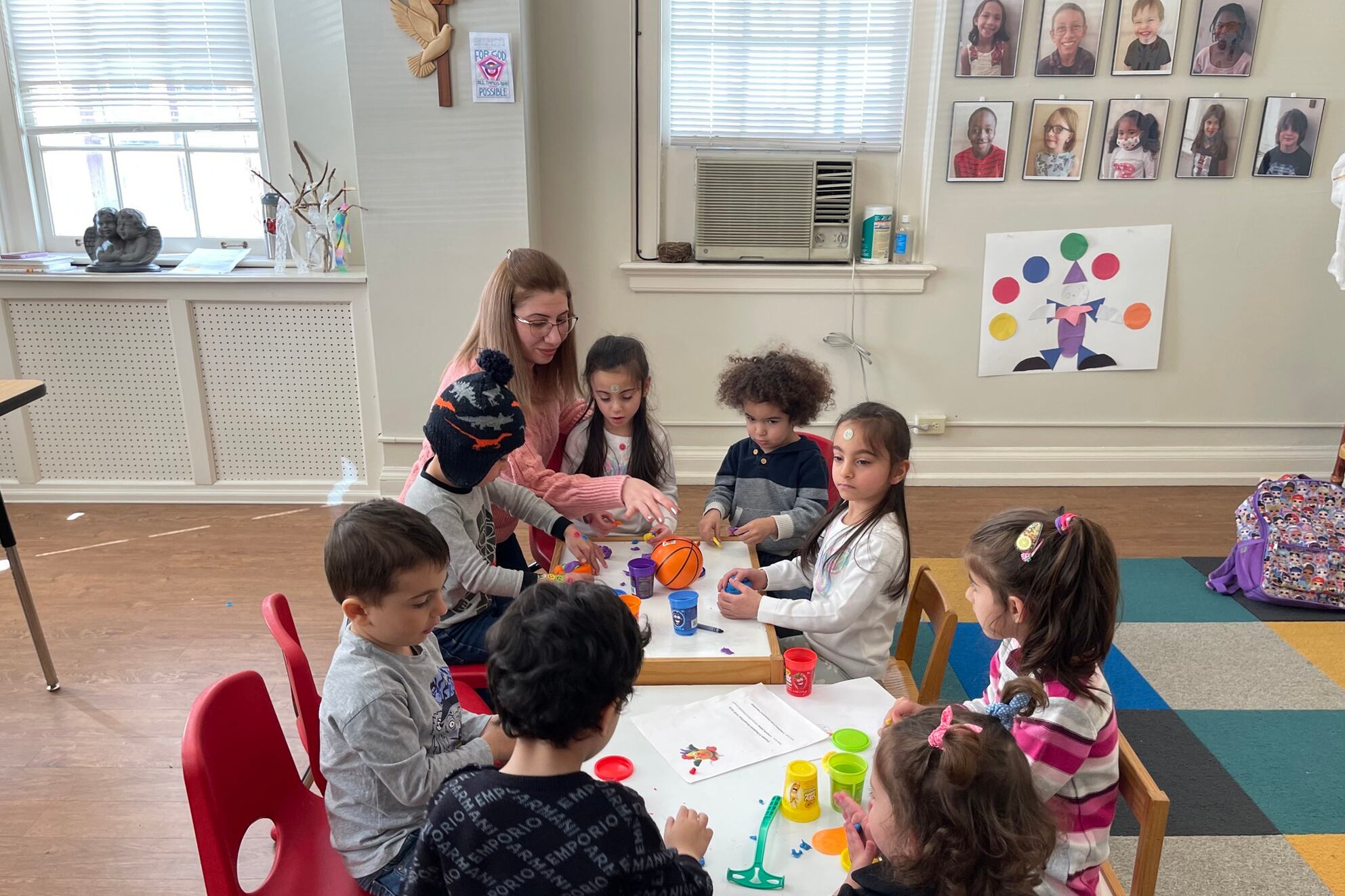 A group of young children sit around a table with an adult in a classroom. There is a colorful rug on the floor and a picture in the background.