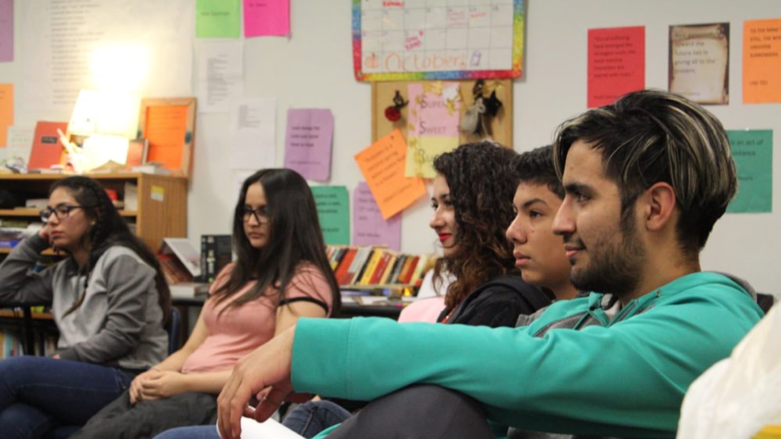 Five students at the New America School in Thornton sit on chairs adjacent to each other in front of a wall with papers and posters on it. 