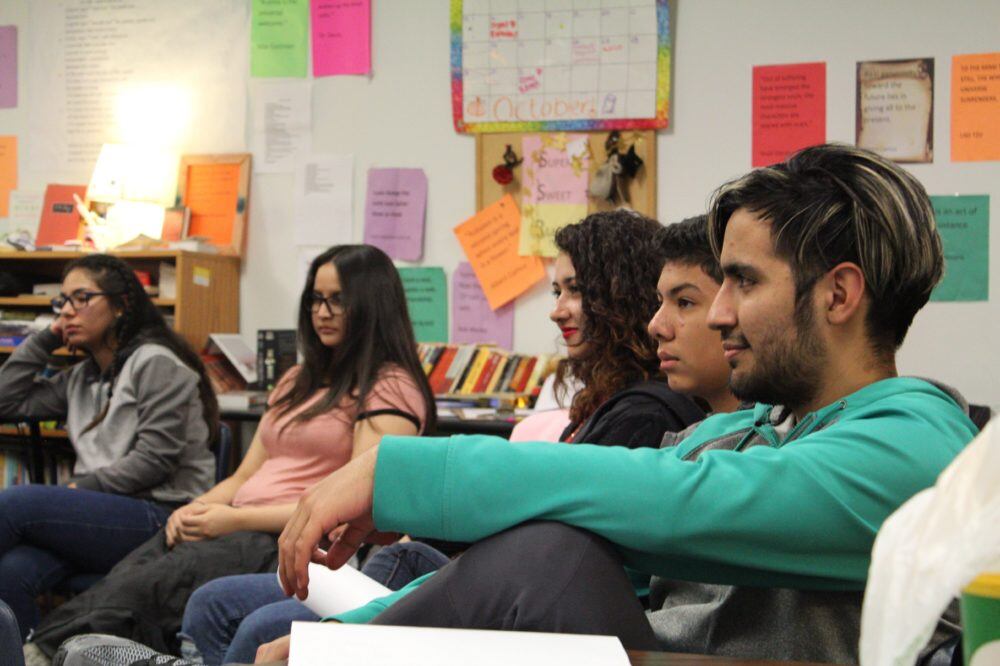 Five students at the New America School in Thornton sit on chairs adjacent to each other in front of a wall with papers and posters on it.