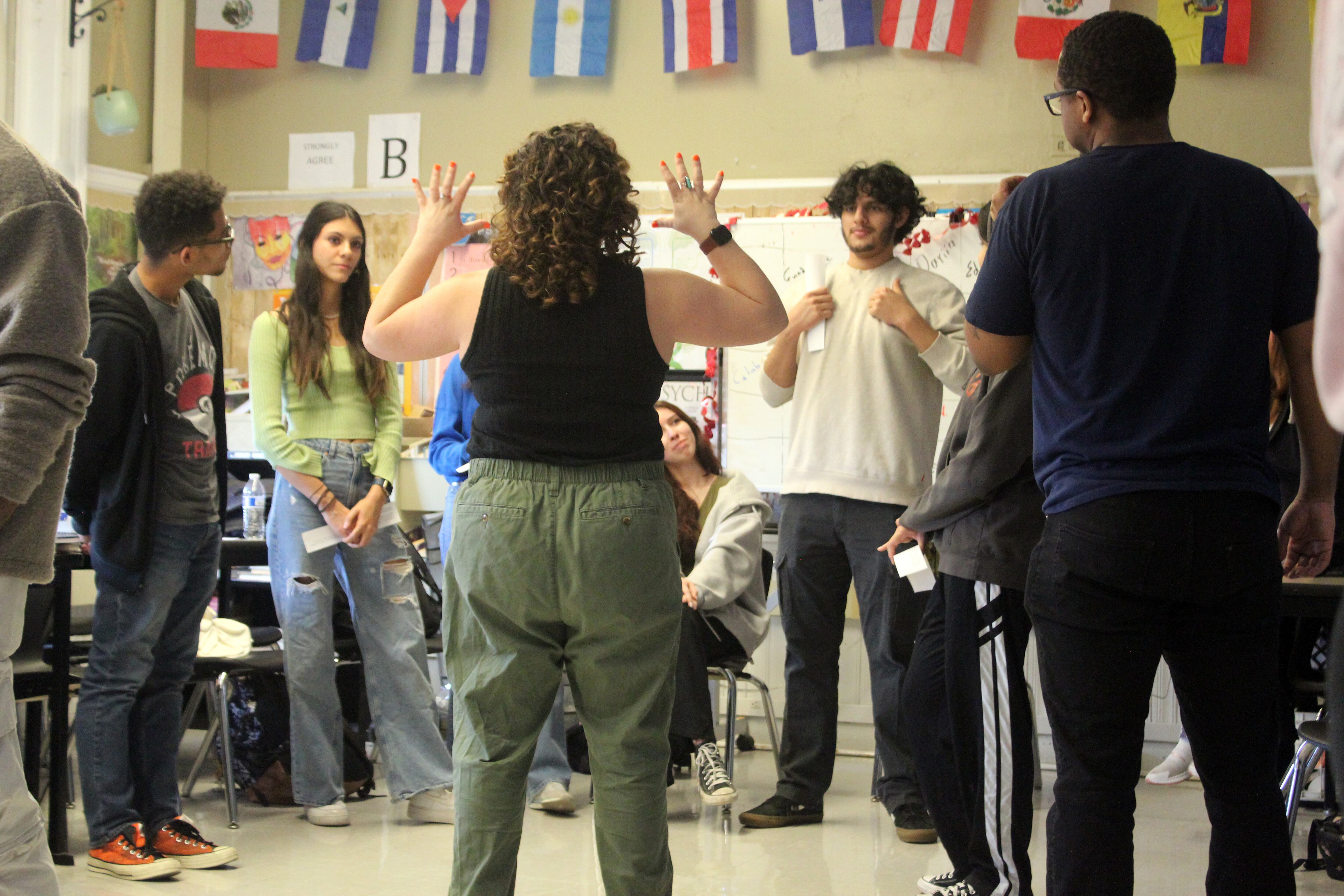 An adult holds her hands up in front of a group of high school students standing in a classroom.