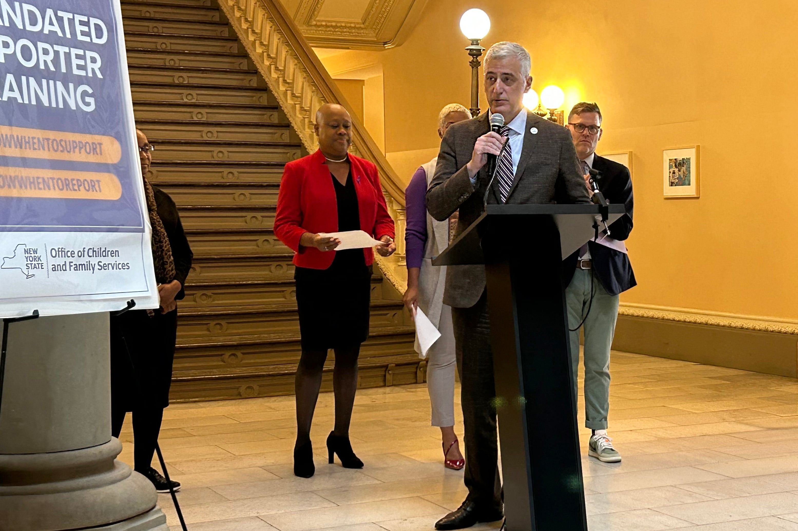 A man in a suit speaks at a podium in front of several other people and a sign that says “Mandated Reporter Training.”