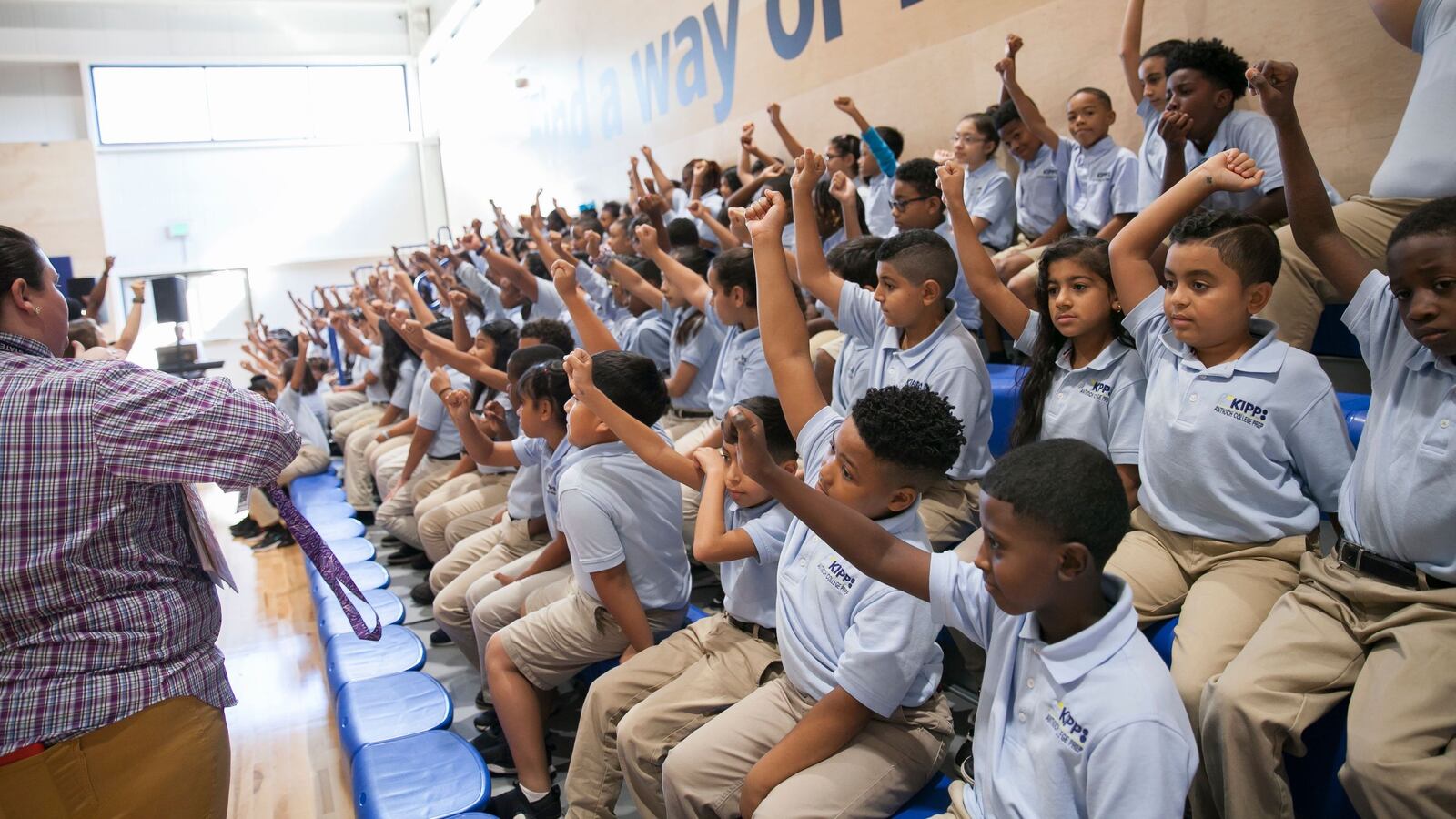A large group of students, wearing light blue shirts and khaki pants, raise their hands seated on bleachers while educators stand in front of them.