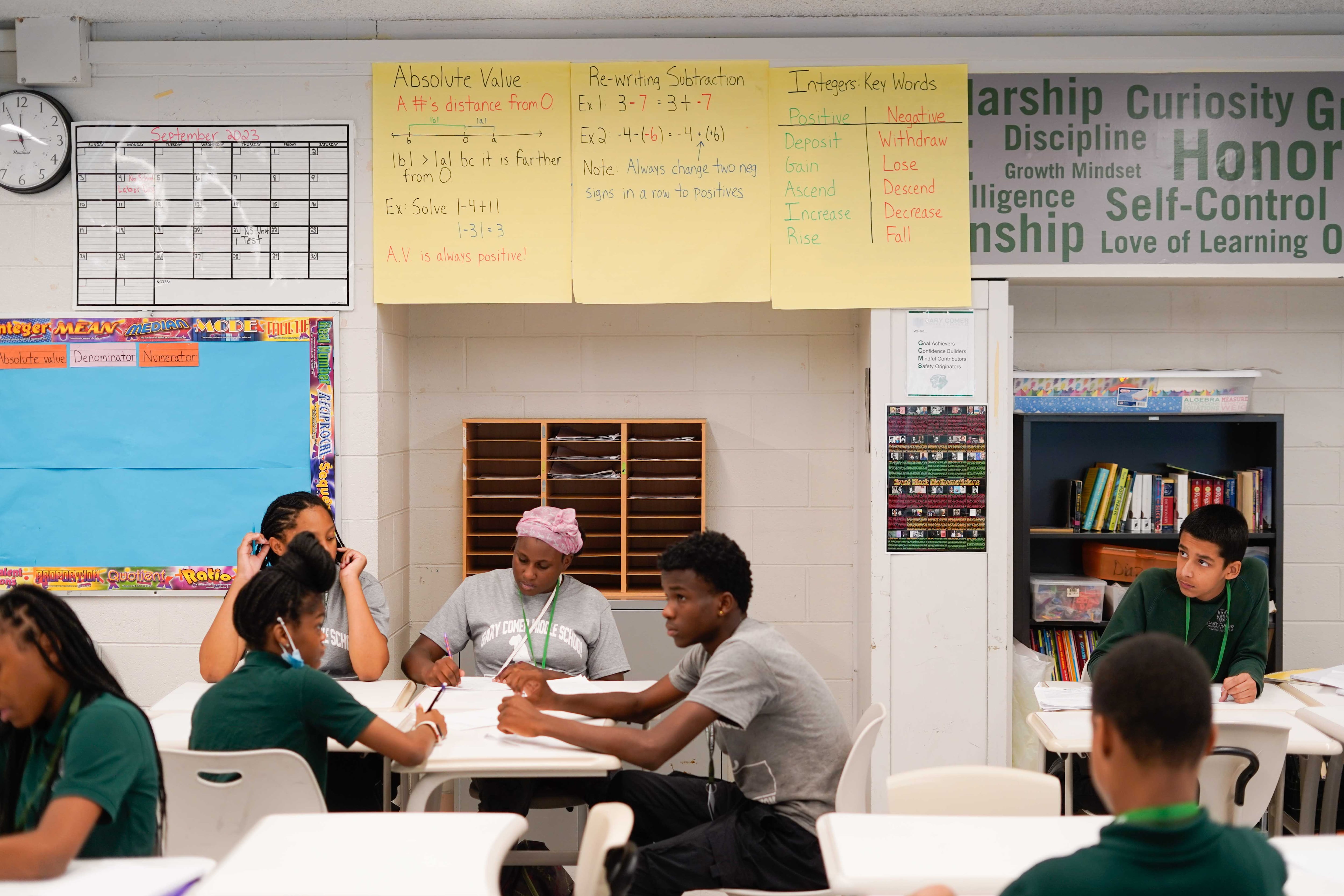 Students sit at their desks with a wall full of posters in the background.