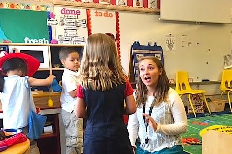 A preschool teacher sitting on the floor talks with one of her students, who is facing her.