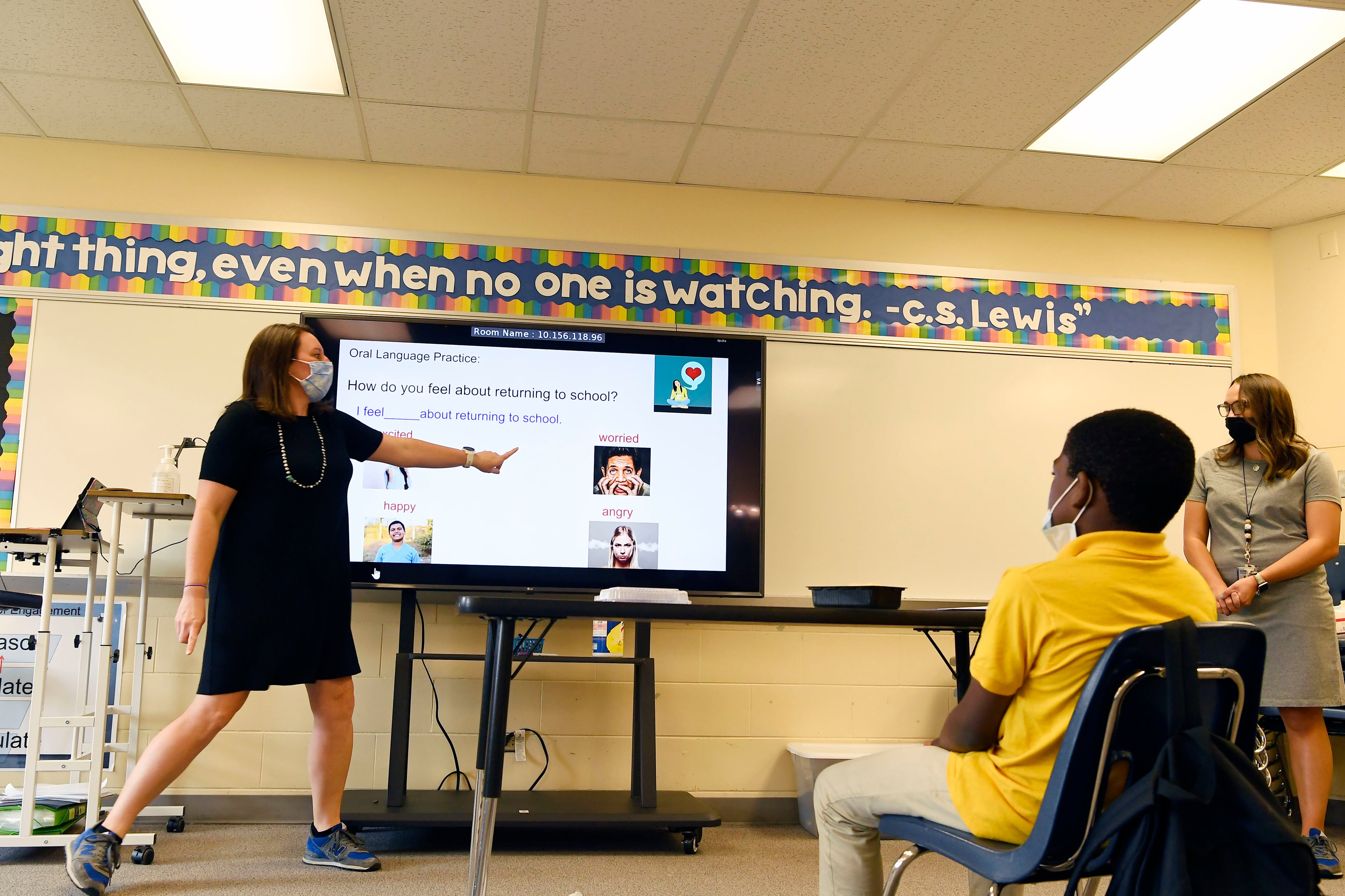 An Aurora teacher wearing a mask points at a screen as students watch.