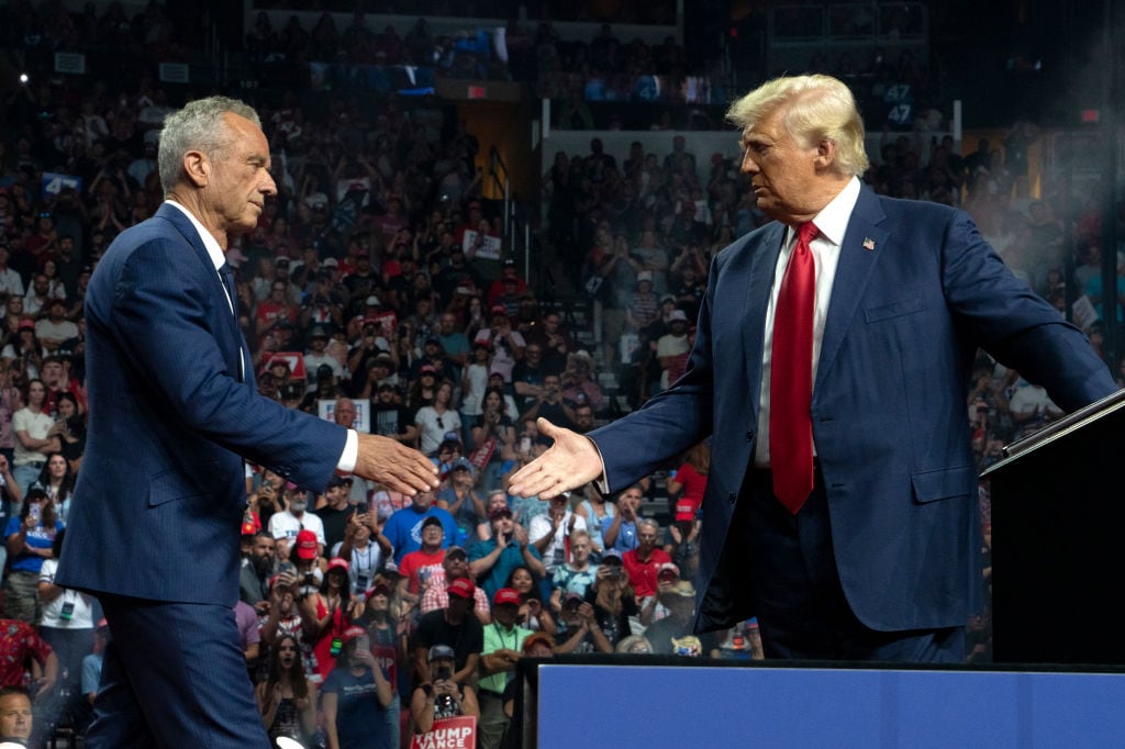 Two older white men in suits extend their hands to shake hands on a rally stage with a large crowd behind them. On the left is former presidential candidate Robert F. Kennedy Jr. On the right is former President Donald Trump.