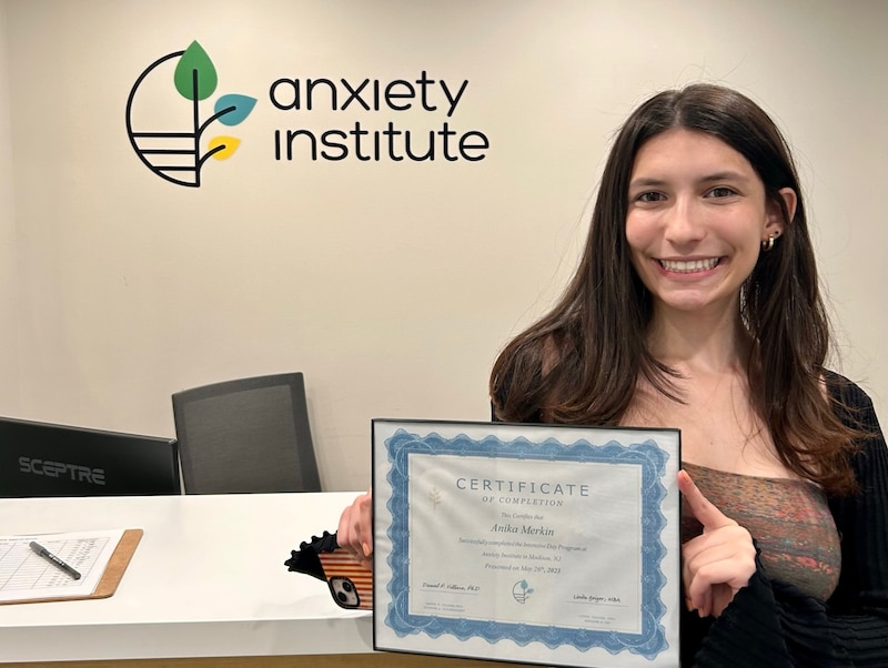 A teenage girl holds up a certificate. In the background, there's a sign that says Anxiety Institute.