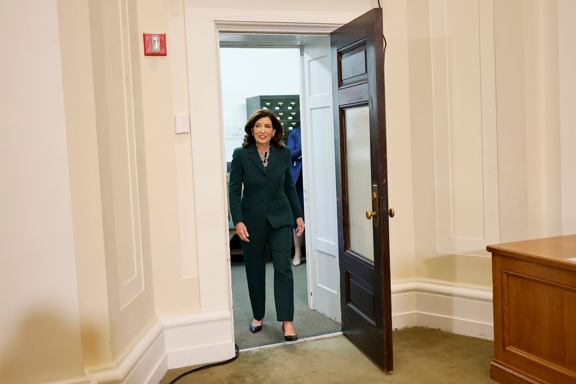 A woman wearing a dark suit walks through a doorway with light tan walls on both sides.