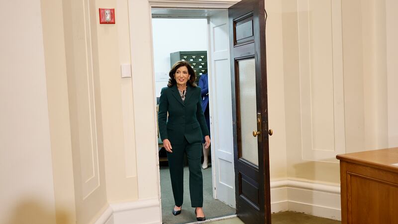 A woman wearing a dark suit walks through a doorway with light tan walls on both sides.