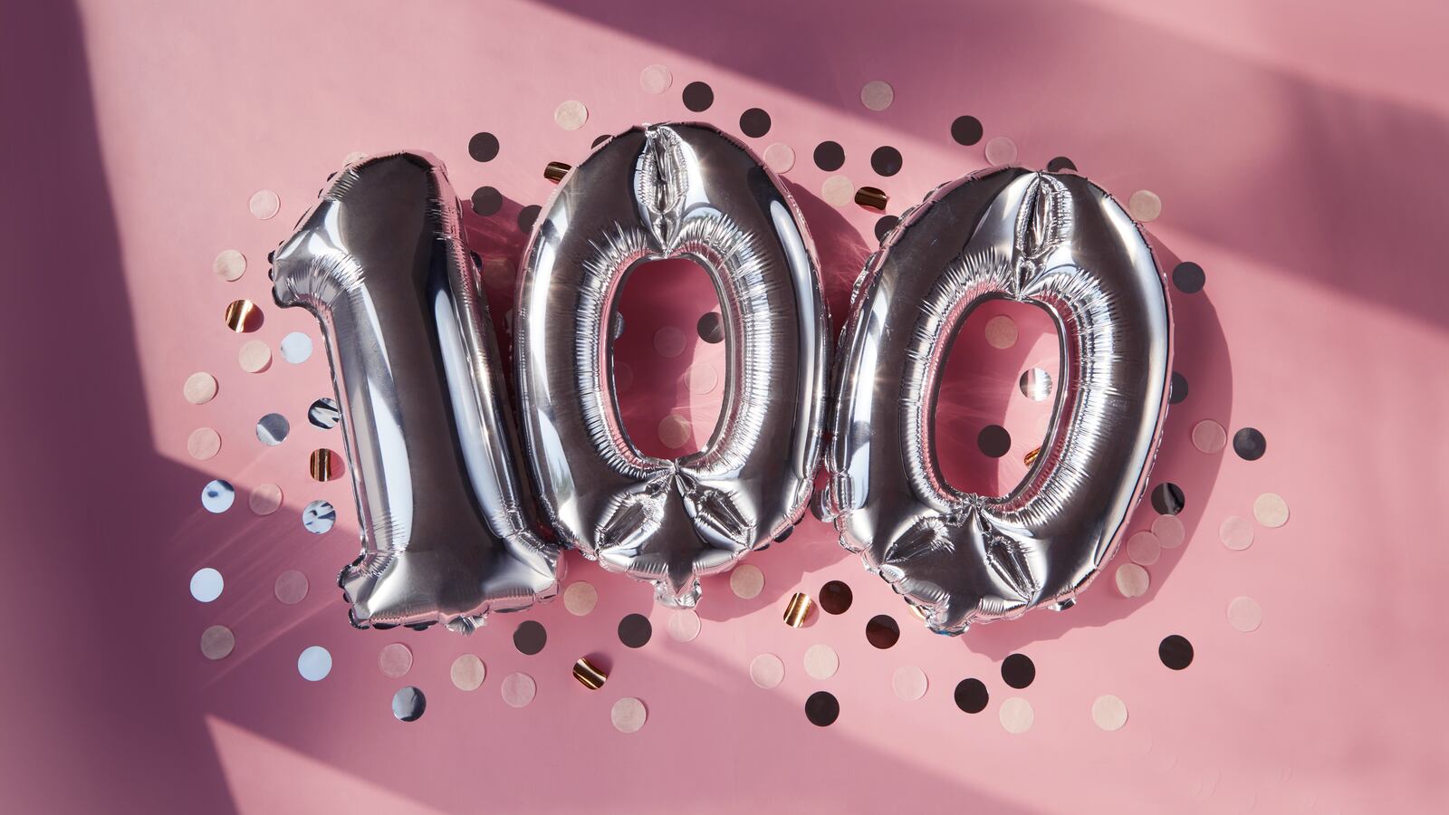 Silver numbers 100 one hundred years balloons among confetti in sunlight on pink purple lilac background.