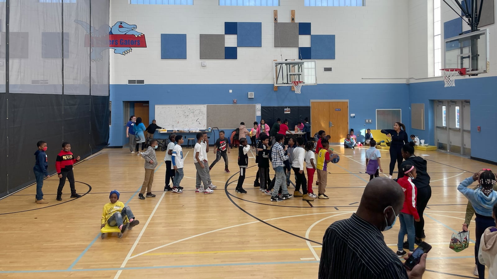 Students in a school gymnasium, lining up in front of a basketball hoop.