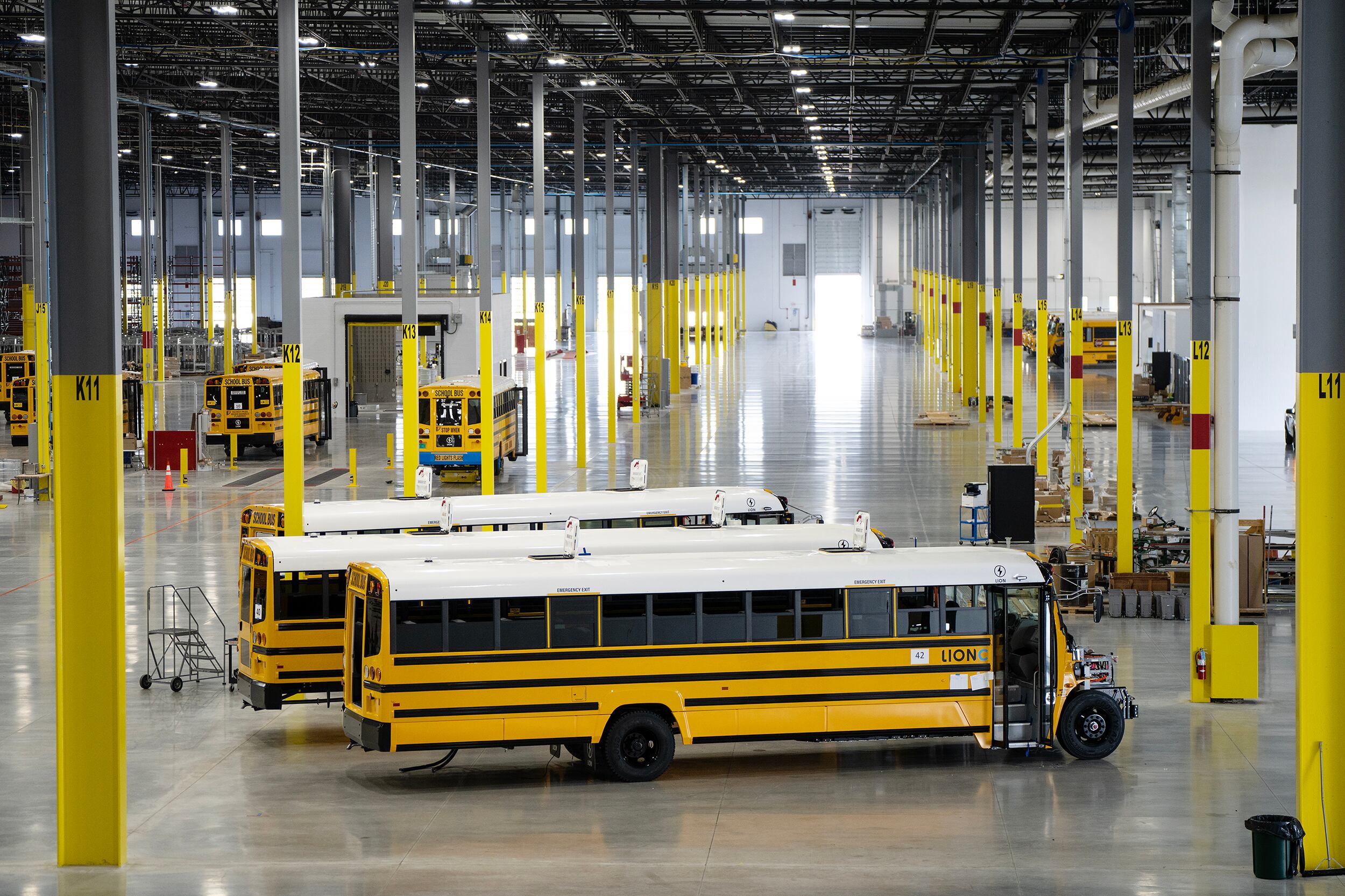 A photograph of three large yellow, white and black school busses lined up in a large warehouse with a couple of busses in the background.