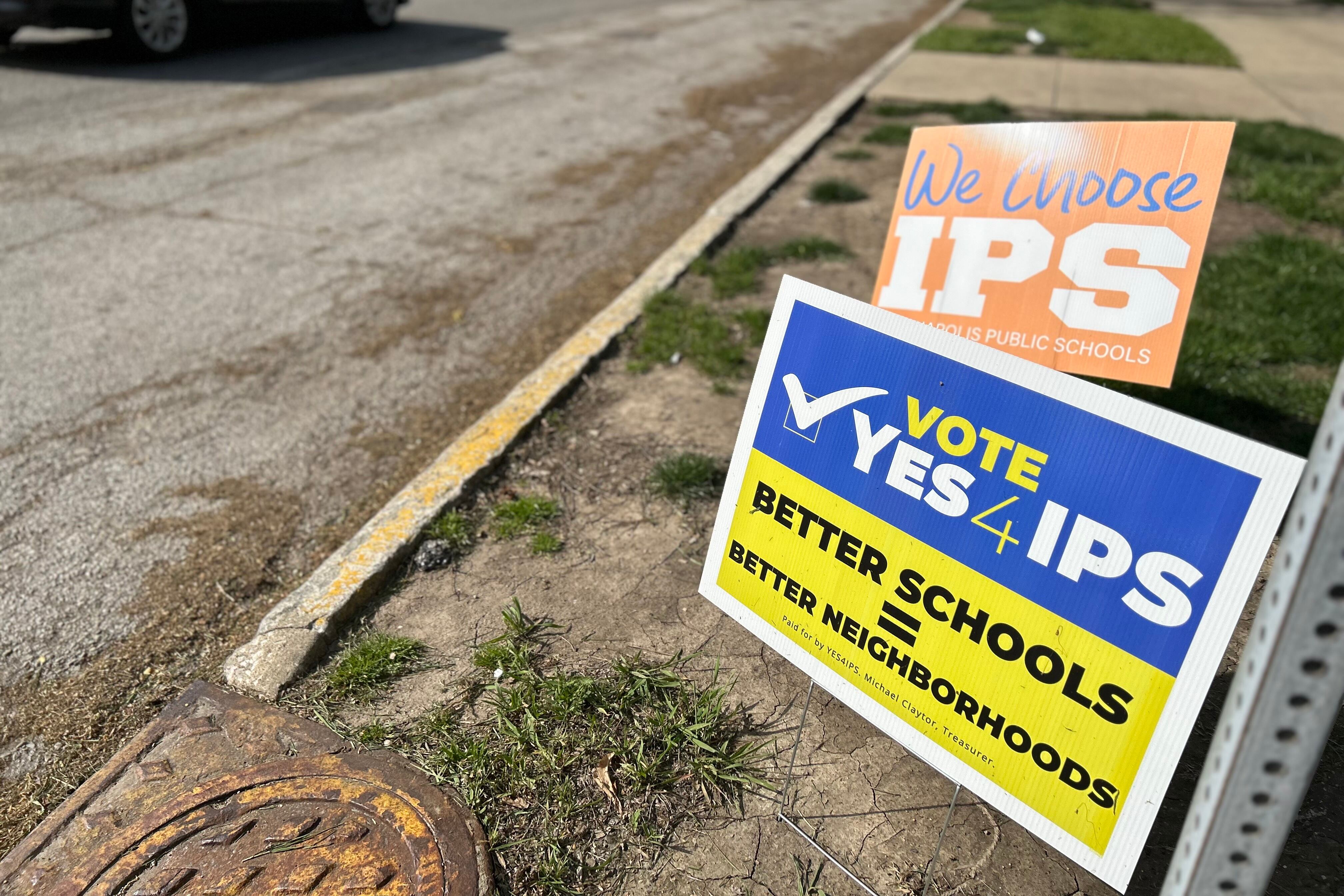 Two signs are in the ground next to teh street. One says “We choose IPS” and the other says “Vote yes for IPS. Better schools equal better neighborhoods.”