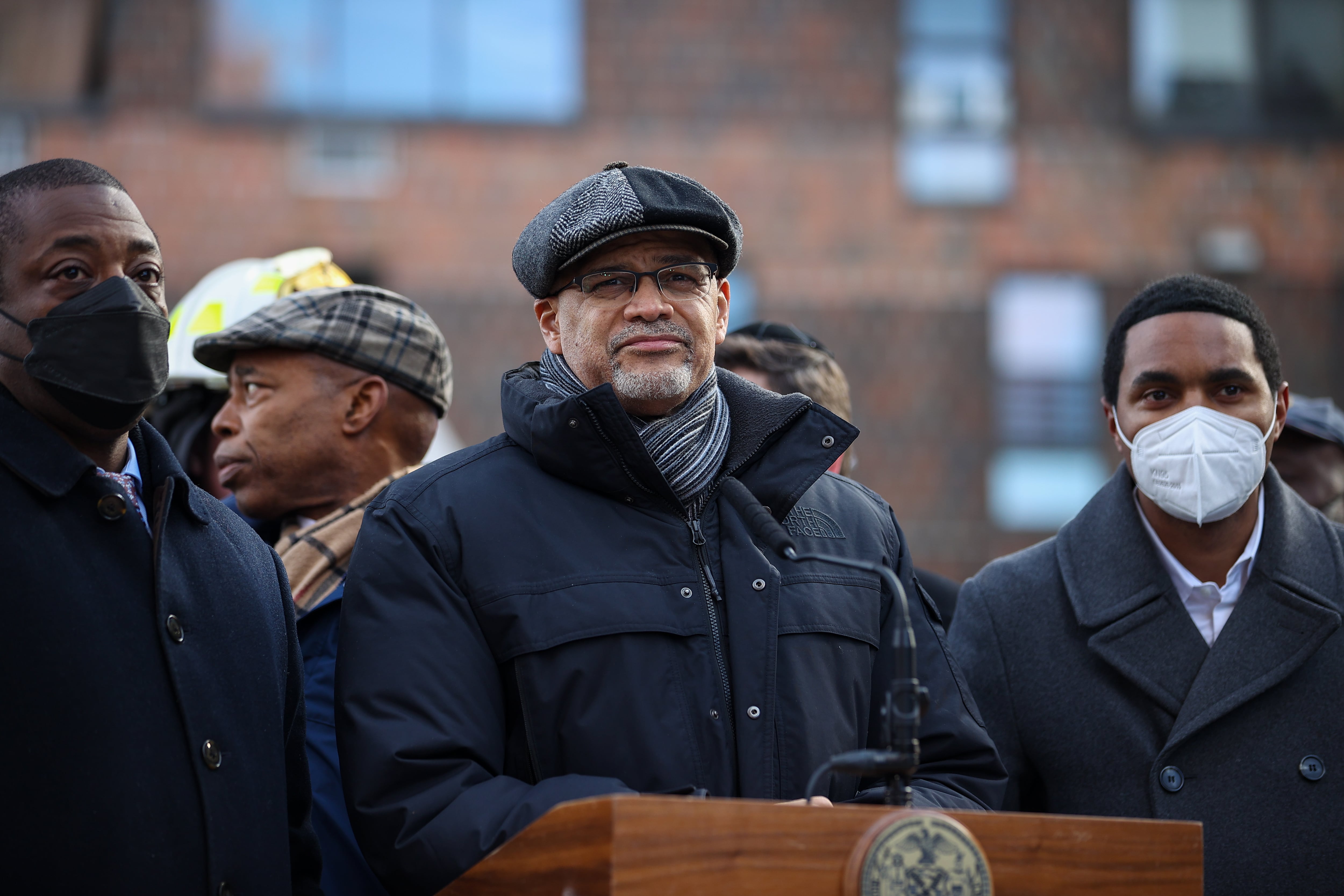A man in winter clothing stands outside at a lectern with other men behind and to each side of him.
